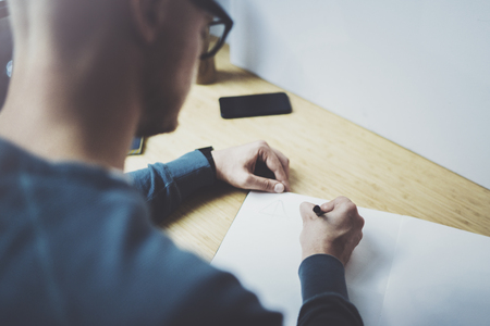 Skilled designer caucasian man drawing abstract sketch with pen.Art work process.Creative hobby.Noting ideas in copy book on workspace desk with papers in modern studio with panoramic windows.Closeup.の写真素材