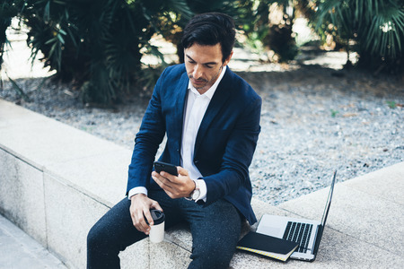 Attractive smiling businessman using contemporary smartphone and laptop while taking rest during work day.Holding hands take away coffee cup.Blurred background. Horizontal.の写真素材