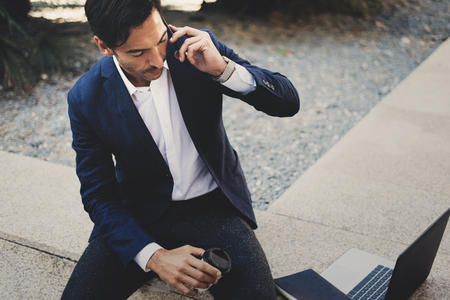 Businessman using contemporary smartphone taking rest outside the office during work day.Holding hands take away coffee cup.Blurred background. Horizontal.の写真素材