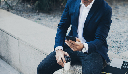 Attractive smiling businessman using contemporary smartphone taking rest during work day.Holding hands take away coffee cup.Blurred background. Horizontal.Cropped.の写真素材