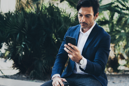 Attractive smiling businessman making video conversation with partners by mobile phone.Man using contemporary smartphone sitting at city park.Blurred background. Horizontal.の写真素材