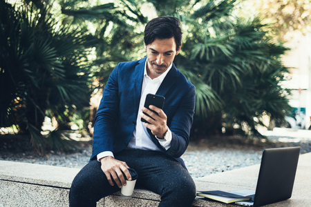 Attractive smiling businessman making video conversation with partners by mobile phone.Man using contemporary smartphone sitting outside.Blurred background. Horizontal.の写真素材