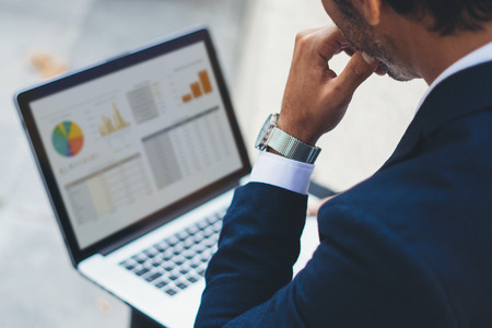 Attractive elegant businessman using contemporary notebook sitting outside the office.Graphs and diagramm on laptop screen.Blurred background. Horizontal.Cropped.の写真素材