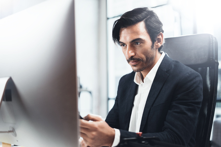 Young elegant man working at sunny office on desktop computer while sitting at wooden table.Blurred background,horizontal.の写真素材