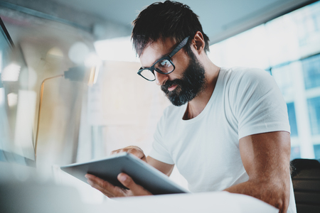 Bearded man in white tshirt wearing eye glasses and using portable electronic pro tablet computer at modern lightful office.Horizontal.Blurred background.の写真素材