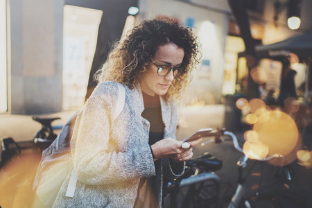 Female hipster wearing backpack and glasses searching information in mobile network by smart phone, during walk in european city at the night. Bokeh and flares effects on blurred background.の写真素材
