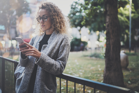 Female hipster wearing backpack and glasses searching information in mobile network by smart phone, during walk in european city park. Bokeh and flares effects on blurred background.の写真素材
