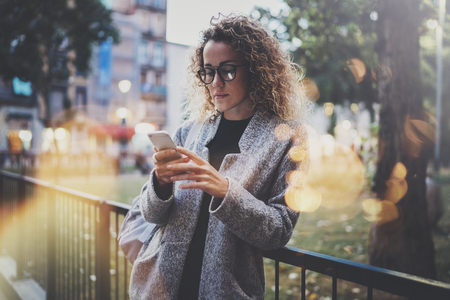 Female hipster wearing backpack and glasses searching information in mobile network by smart phone, during walk in european city park. Bokeh and flares effects on blurred background.の写真素材