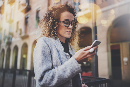 Hansome girl hipster wearing glasses searching information in mobile network by smart phone, during walk in european city. Bokeh and flares effects on blurred background.の写真素材