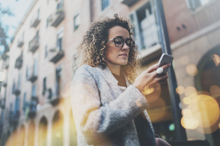 Hansome girl hipster wearing glasses searching information in mobile network by smart phone, during walk in european city. Bokeh and flares effects on blurred background.の写真素材