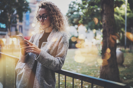 Female hipster wearing backpack and glasses searching information in mobile network by smart phone, during walk in european city park. Bokeh and flares effects on blurred background.の写真素材
