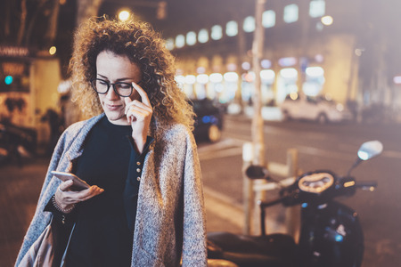 Beautiful girl hipster wearing glasses holding smart phone hands, during night walking in european vocation. Bokeh and flares effects on blurred background.の写真素材