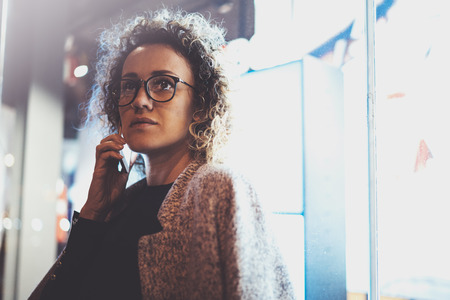 Pensive pretty woman making conversation call with her friends via cell telephone while standing at night on the street. Bokeh and flares effect on blurred background.の写真素材