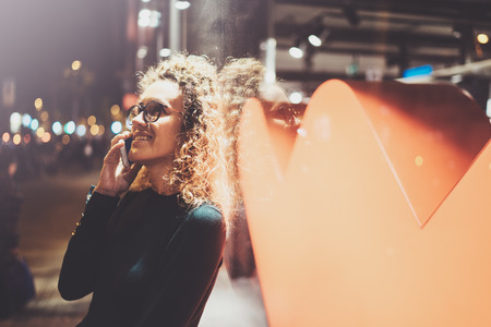 Smiling pretty woman making conversation call with her friends via cell telephone while standing at night on the street. Bokeh and flares effect on blurred background.の写真素材