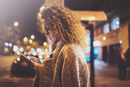 Beautiful girl hipster wearing glasses holding smart phone hands, during night walking in european vocation. Bokeh and flares effects on blurred background.の写真素材