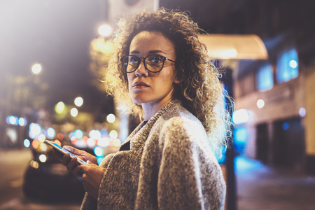 Beautiful girl hipster wearing glasses holding smart phone hands, during night walking in european vocation. Bokeh and flares effects on blurred background.の写真素材
