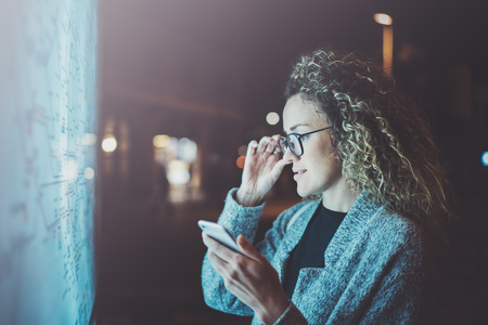 Charming fashionable woman with eyes glasses looking on screen smartphone on background light box in night atmospheric city map. Bokeh and flares effect on blurred background.の写真素材