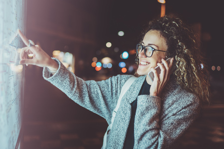 Happy fashionable woman tourist with eyes glasses talking on mobile phone while looks at map of the city on electronic bulletin board. Bokeh and flares effect on blurred background.の写真素材