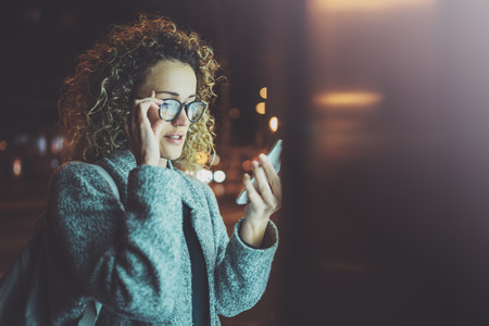 Woman in eye glasses holding hands smartphone in night atmospheric city.Female hands using mobile phone.Closeup on blurred background.Flares, bokeh effects.の写真素材