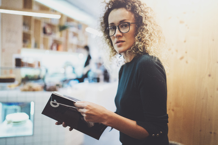 Charming fashionable woman with eyes glasses reading magazine sitting indoor in urban cafe.Casual portrait of pretty girl. Blurred background.の写真素材