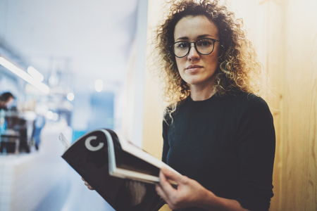 Charming fashionable woman with eyes glasses reading magazine sitting indoor in urban cafe.Casual portrait of pretty girl. Blurred background.の写真素材