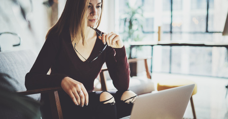 Young beautiful blonde woman sitting at the comfortable armchair and using laptop computer.Working process at coworking studio.Panoramic windows on blurred background.の写真素材