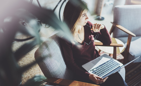 Young beautiful blonde woman sitting at the comfortable armchair and using laptop computer.Working process at coworking studio.Panoramic windows on blurred background.の写真素材