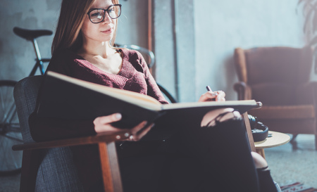 Handsome young girl wearing glasses casual clothes holding book hands.Woman blonde sitting in vintage armchair modern loft studio reading book. Blurred background.Horizontal.の写真素材