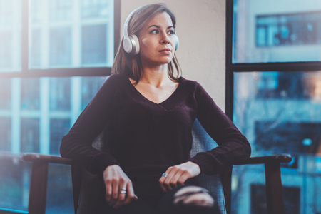 Beautiful blonde girl wearing eye glasses and casual clothes listening digital music in headphones on smartphone while relaxing in armchair at modern place.の写真素材