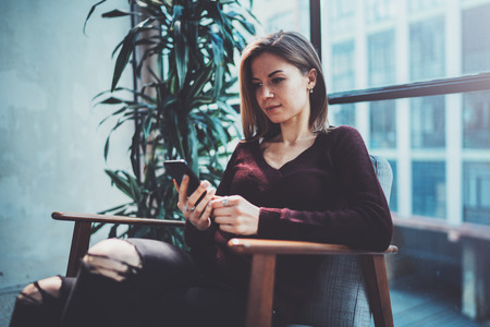 Blonde yound woman holding modern mobile phone in hands.Girl pointing fingers on empty touch mobile screen at sunny coworking place. Horizontal, blurred background.の写真素材