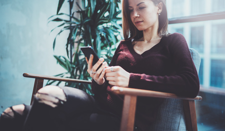 Blonde yound woman holding modern mobile phone in hands.Girl pointing fingers on empty touch mobile screen at sunny coworking place. Horizontal, blurred background.の写真素材