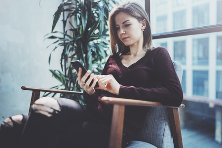 Blonde yound woman holding modern mobile phone in hands.Girl pointing fingers on empty touch mobile screen at sunny coworking place. Horizontal, blurred background.の写真素材