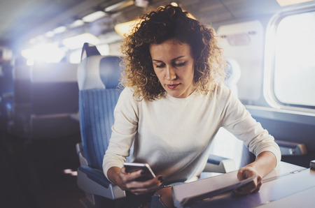 Enjoying travel concept. Young pretty woman tourist traveling by the train sitting near the window using smartphone.の写真素材