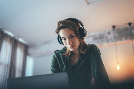 Young handsome girl relaxing at modern home,wearing casual clothes,playing music on smartphone and listening audio books in wireless headphones.Blurred background.の写真素材