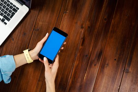 Mockup image of a woman using mobile phone with blank screen on wooden table. Close up photo of female hands holding smartphone uprightの写真素材