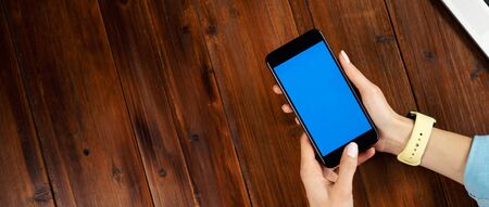 Mockup image of a woman using mobile phone with blank screen on wooden table. Close up photo of female hands holding smartphone uprightの写真素材