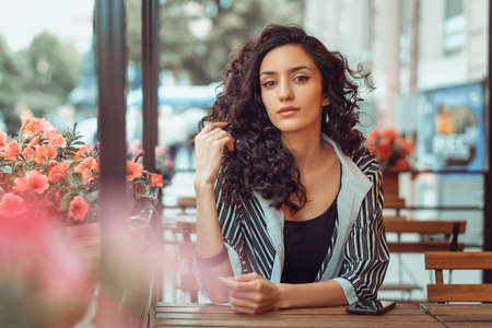 portrait of a beautiful girl with curly hair sitting at a table in a cafe on the street.の写真素材