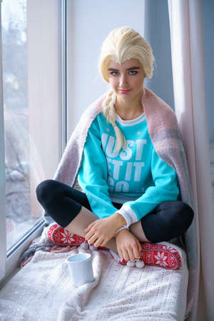 a beautiful blonde girl in a blue sweater and new year's slippers sits on the windowsill with a mug...