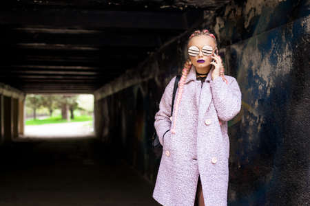 A fashionable woman with pink pigtails in a pink coat with a bright make up and round glasses poses on the street in a tunnel.の写真素材