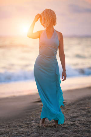 A young slender girl stands on the beach at sunset in a blue long dress, a beautiful figure.の写真素材