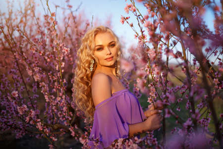 Beautiful young blonde woman with wavy hair posing with blooming peach trees against the sky.