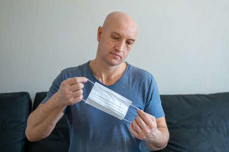 Portrait of a bald man sitting at home on the sofa holding a medical mask in his hands.