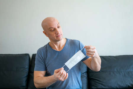 Portrait of a bald man sitting at home on the sofa holding a medical mask in his hands.の写真素材
