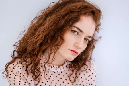 A young red-haired girl with blue eyes, dressed in a blouse with polka dots, looks at the camera with a serious look.の写真素材
