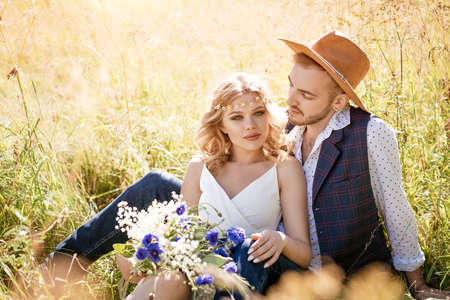 A young man in a hat and a beautiful girl with a make-up and hairstyle, hugging sitting in a field in the grass on a Sunny day.の写真素材