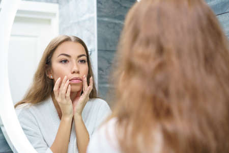 Beautiful young woman looks at her reflection in the mirror and gives a facial massage.の写真素材