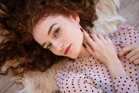 A young red-haired girl with blue eyes, dressed in a dress with polka dots, lies on a carpet made of...