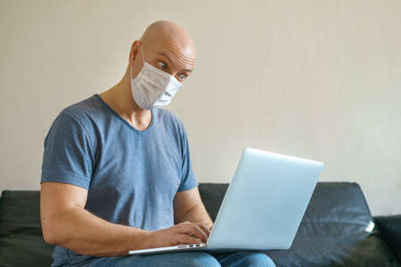 A bald man works at home at a laptop on a sofa in a mask during quarantine. The concept of remote work.の写真素材