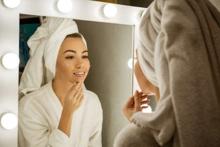 A happy young woman in a towel in front of a mirror applies cream to her face, a concept of skin care at home.の写真素材