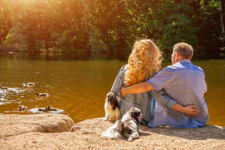 Happy Mature couple sitting on the lake in the sun with their dogs. Concept of family vacation in nature.の写真素材